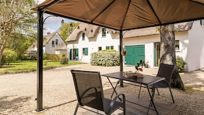 Een overdekt buitengebied met een tafel, stoelen, een boom en een gebouw op de achtergrond.