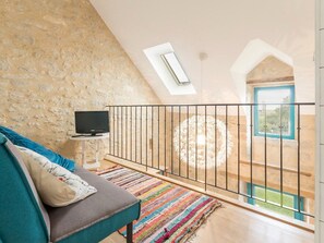 Une pièce chaleureuse avec un mur en pierre, un téléviseur posé sur une petite table, un tapis rayé aux couleurs vives et un balcon doté d’une rambarde en métal, offrant une vue sur la verdure.