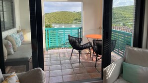 Un balcón con una mesa y sillas, vista a barcos en el agua y una colina verde al fondo.