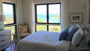 Un dormitorio con una cama grande, una silla, un aparador y vistas al mar a través de amplias ventanas.