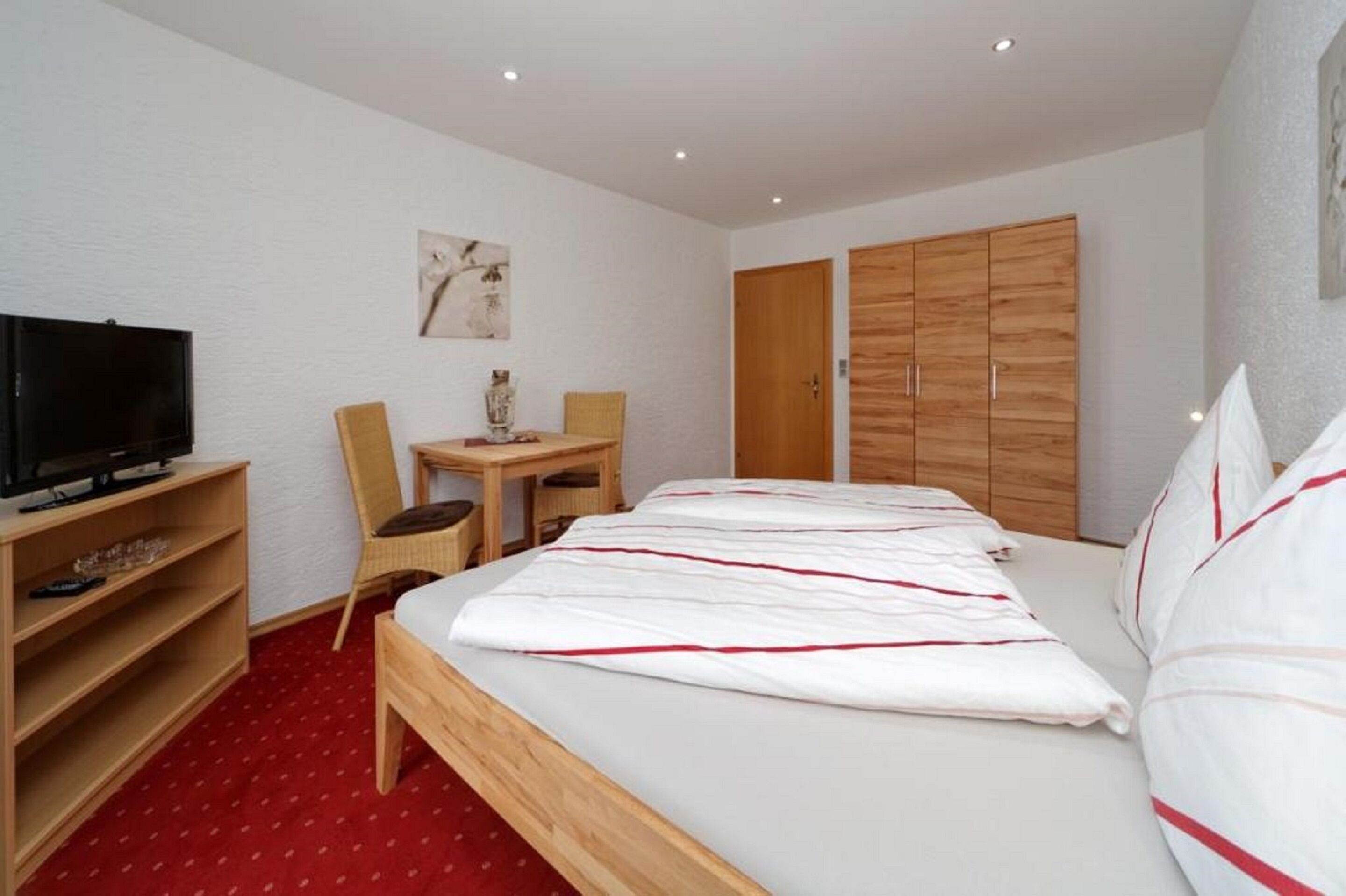 A bedroom with a bed, a television on a stand, a wooden wardrobe, and a small table with a chair.