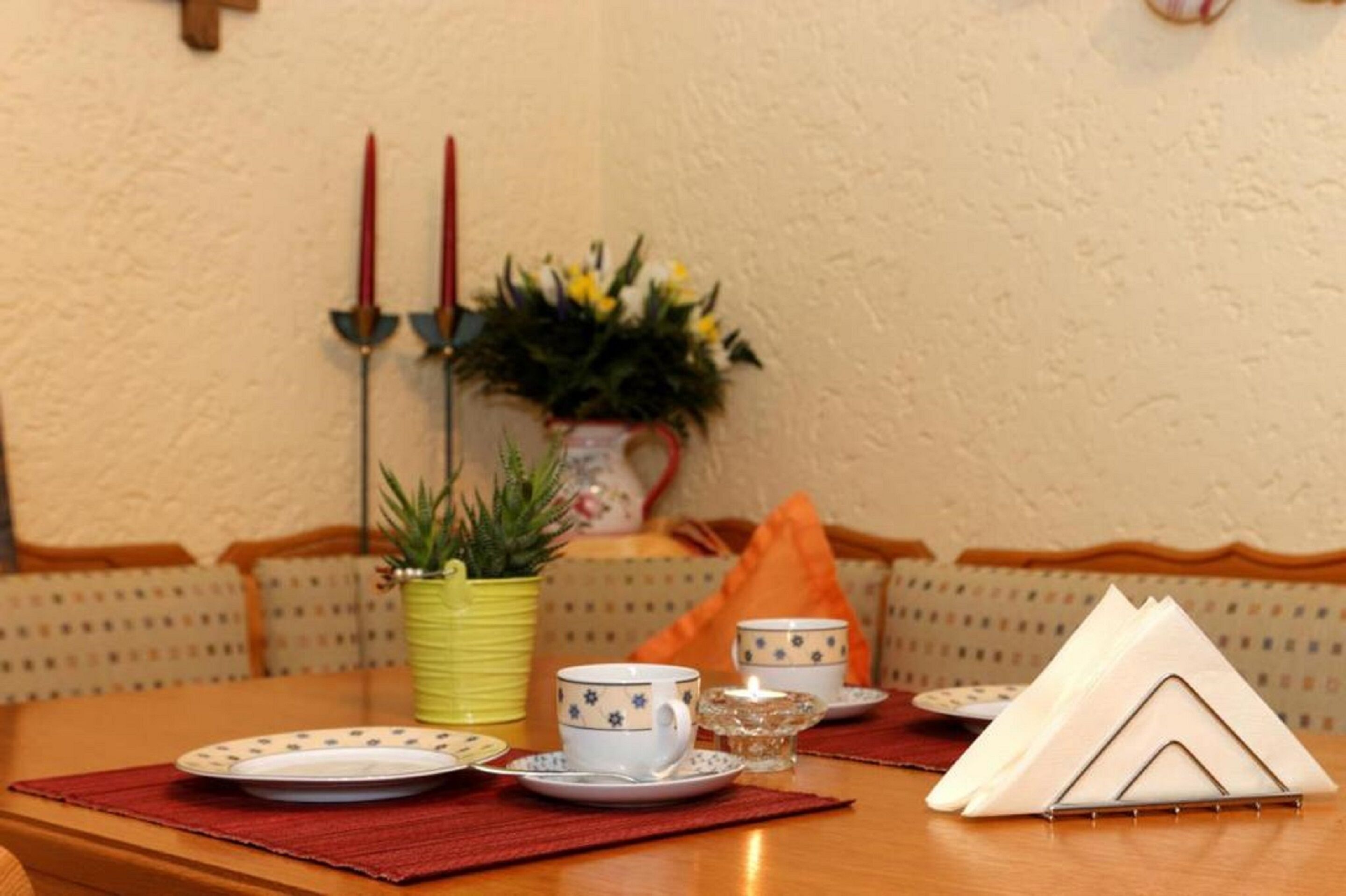 Una mesa de comedor con platos, tazas y una vela. Hay una planta en una maceta amarilla y una pequeña estructura de madera sobre la mesa.