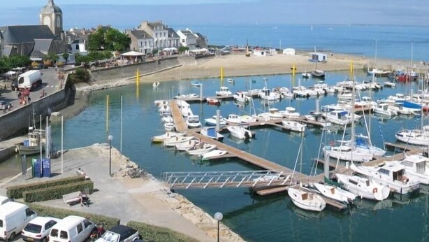 A marina with multiple boats docked, a sandy beach, and buildings along the waterfront.