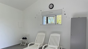 A room with two white lounge chairs, a small table, a clock, and a window with a view of trees.
