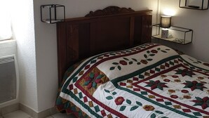 A bedroom with a wooden headboard, a quilted bedspread, a wall-mounted nightstand with a lamp, and a window with blinds.