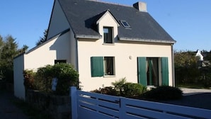 Une maison à deux étages, avec un toit en pente, une petite fenêtre de grenier et des volets verts.