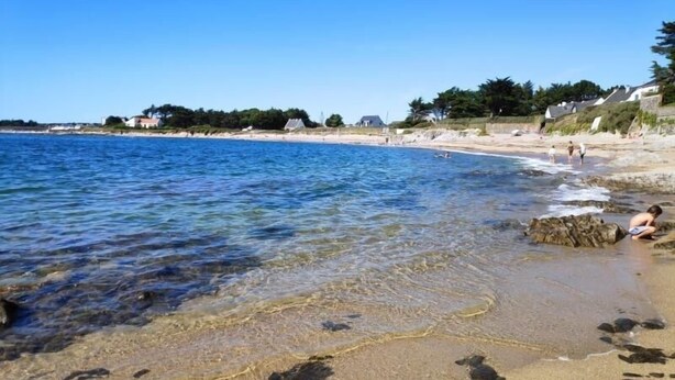 Une plage avec une eau claire, un rivage sablonneux et des maisons en arrière-plan.