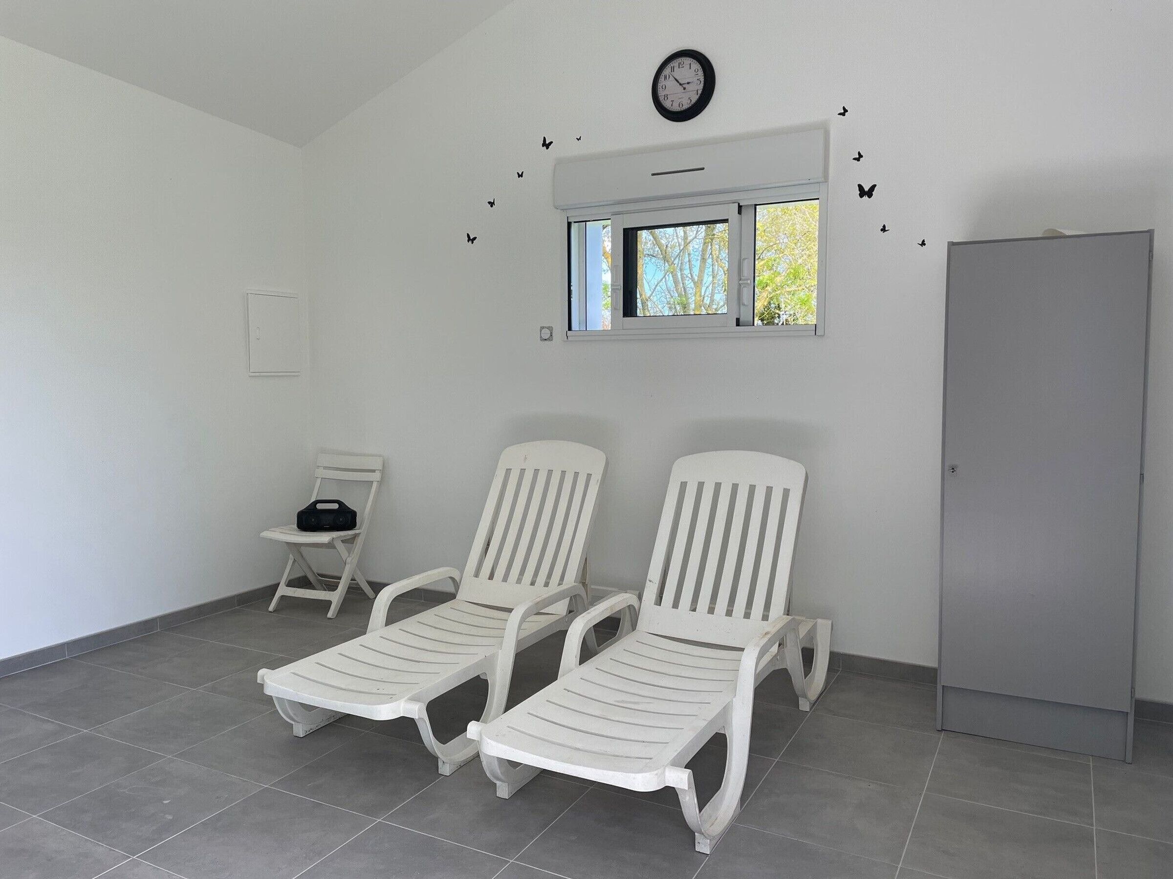 Une pièce comprenant deux fauteuils blancs, une petite table, une horloge et une fenêtre donnant sur des arbres.