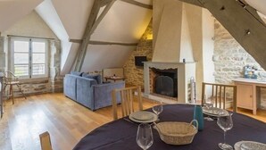 Un espacio tipo loft con vigas de madera a la vista, pared de piedra, chimenea, sofá y mesa de comedor preparada para seis personas.
