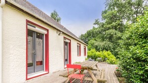 A wooden deck with a red chair and a table, leading to a building with red-framed windows and a white exterior. - Spacious Ground-Level Home with Large Yard, Terrace, WiFi, and Game Amenities in Malville (Malville)