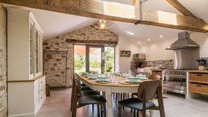 Un espace repas avec une longue table dressée pour six personnes, entourée de chaises, dans une pièce aux poutres apparentes et aux murs en pierre.
