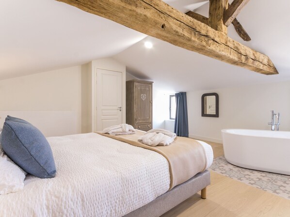 A bedroom with a large bed, a wooden ceiling beam, a wardrobe, a bathroom with a freestanding bathtub, and a window with curtains. - Charming Countryside Retreat with Indoor Heated Pool Near Nantes & Clisson (Château-Thébaud)