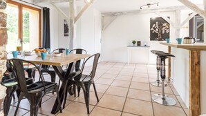 Un coin repas avec une table en bois et des chaises noires, un espace cuisine avec des tabourets de bar, et une vue sur l’extérieur par une fenêtre.