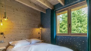 Quarto com teto de madeira, cama com roupa de cama branca, mesa de cabeceira, luminária na parede e janela com cortinas.