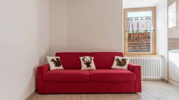 A red sofa with three decorative pillows, a window with a view of a wooden structure, and a white radiator.