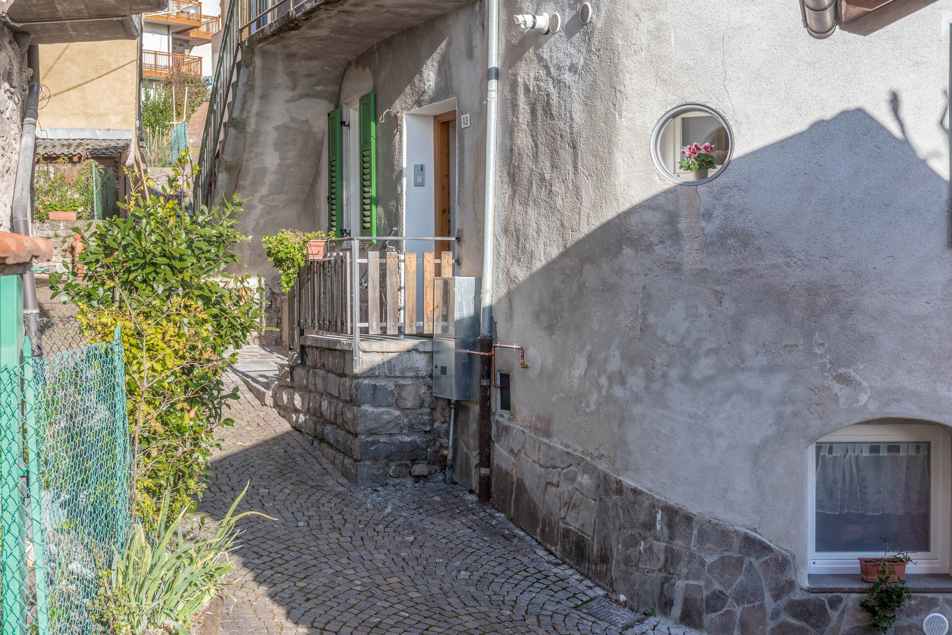 A narrow cobblestone street with a building featuring a stone facade, a small window with a potted plant, and a visible door.