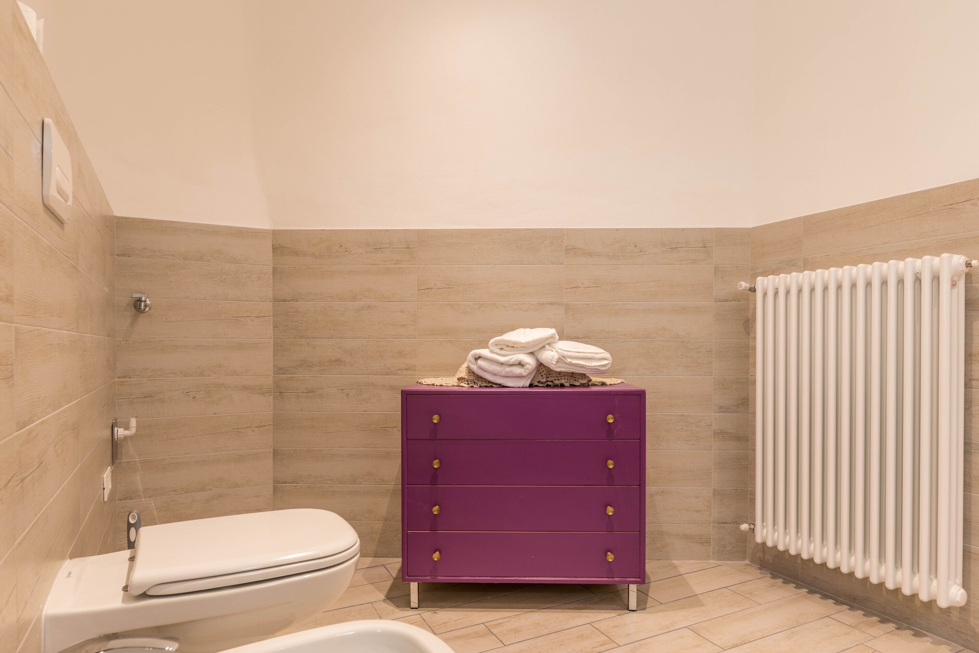 Une salle de bain avec des toilettes blanches, un meuble-lavabo violet doté de tiroirs et un radiateur blanc.