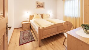 A bedroom with a wooden bed, bedside tables, a nightstand, a desk, a chair, a rug, and a window with curtains. - Holiday Apartment "Désirée Dolasilla" with Balcony, Mountain View, Wi-Fi & TV (St. Vigil)