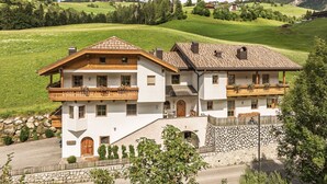 A two-story chalet with a stone wall, wooden balcony, and a view of green hills. - Holiday Apartment "Désirée Dolasilla" with Balcony, Mountain View, Wi-Fi & TV (St. Vigil)