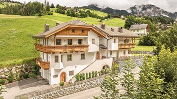 Un chalet à deux étages doté d’un balcon, d’un escalier en pierre et offrant une vue sur une vallée verdoyante et des montagnes.