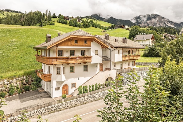 A two-story chalet with a balcony, stone steps, and a view of a green valley and mountains.