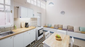 A compact kitchen with white cabinets, a stove, and a sink. There is a wooden table with a bowl of fruit and a white chair. A window provides natural light, and a clock is on the wall. - Home with Terrace in Heart of old Town of Chania (Chania)