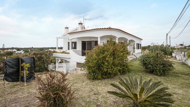 A white house with a curved roof, surrounded by a well-maintained lawn and various shrubs.