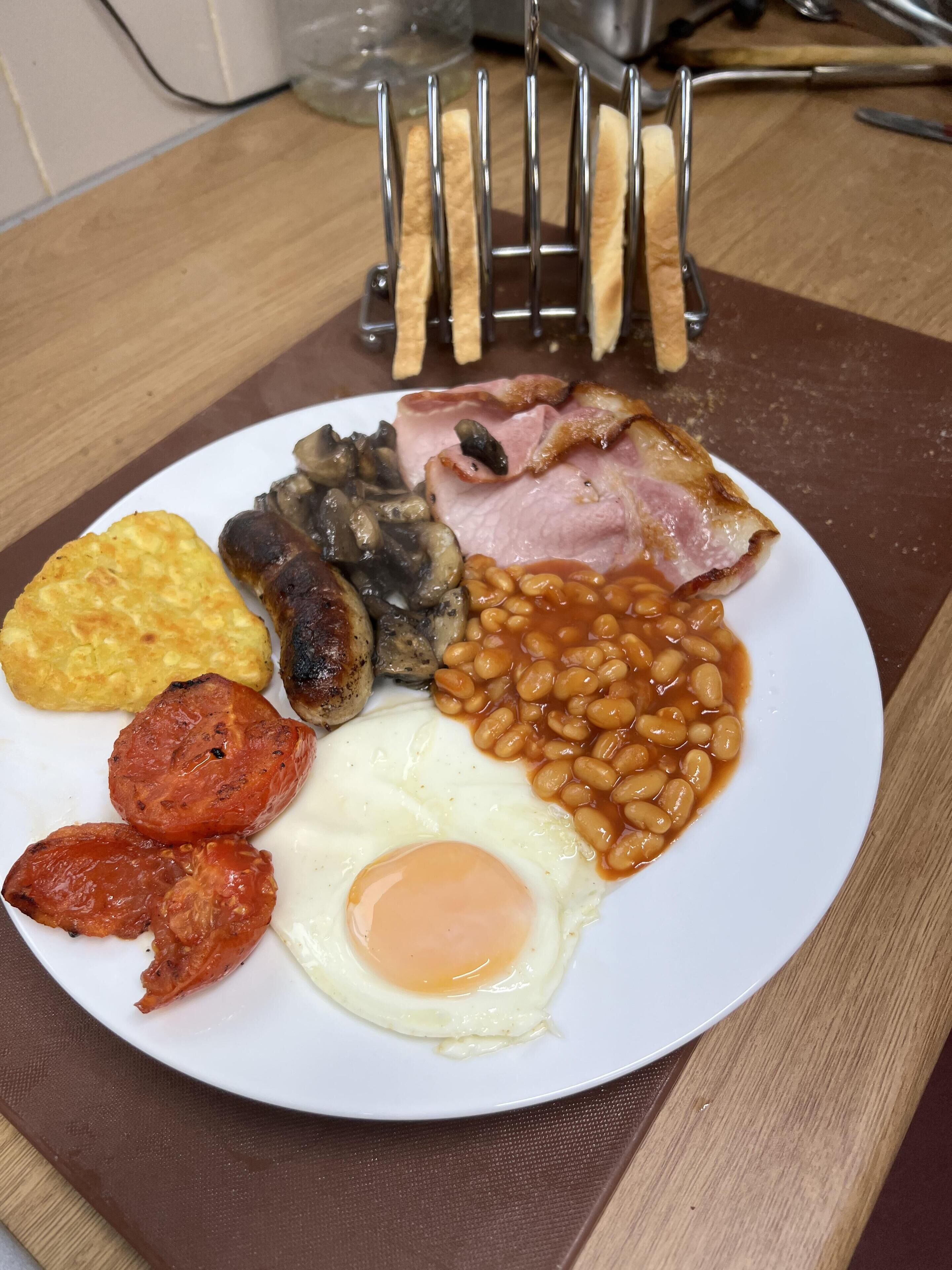 A plate of breakfast food including eggs, sausages, mushrooms, baked beans, and bacon.