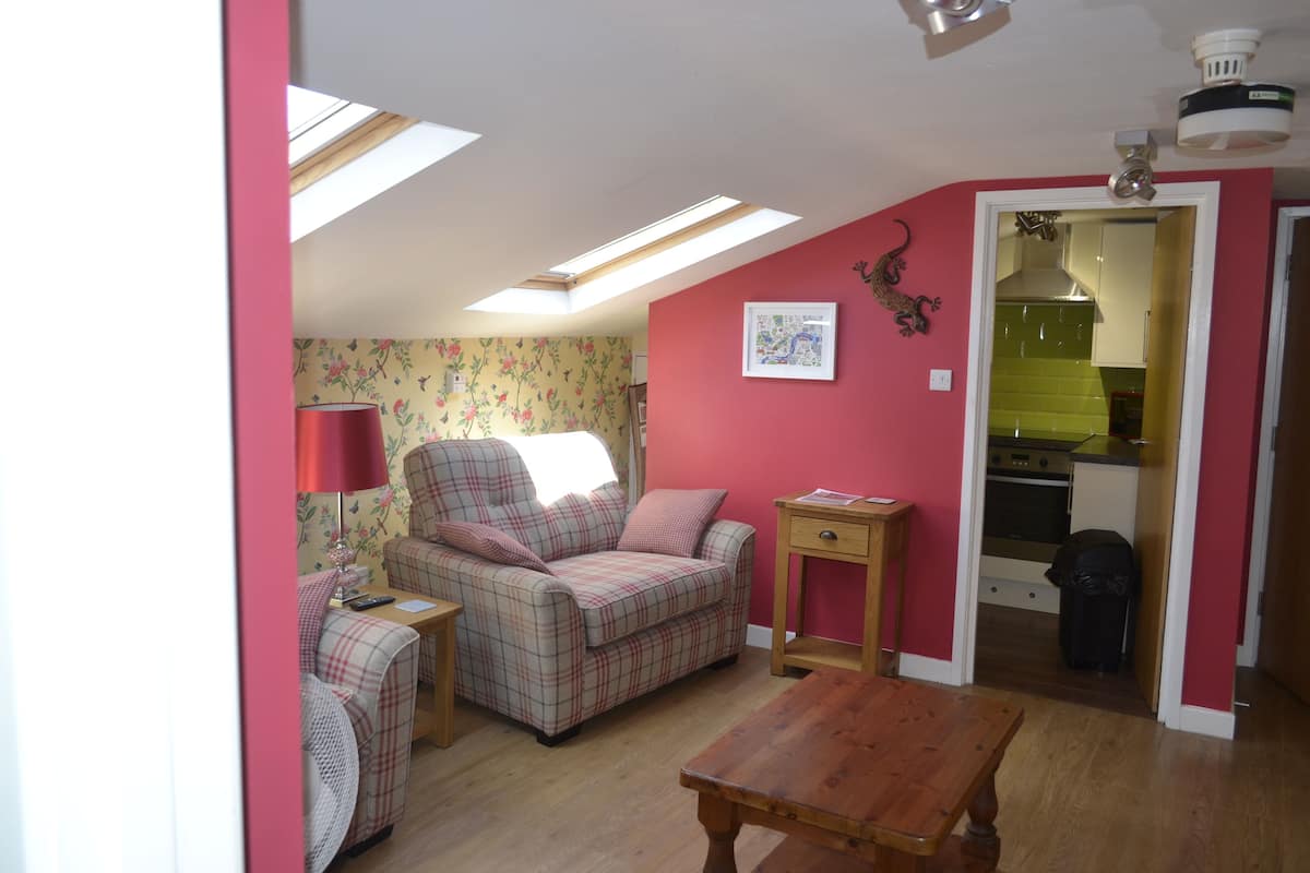 A cozy room with a plaid sofa, a wooden table, and a small side table. There is a window with a view of the outside, and a door leading to a kitchen.