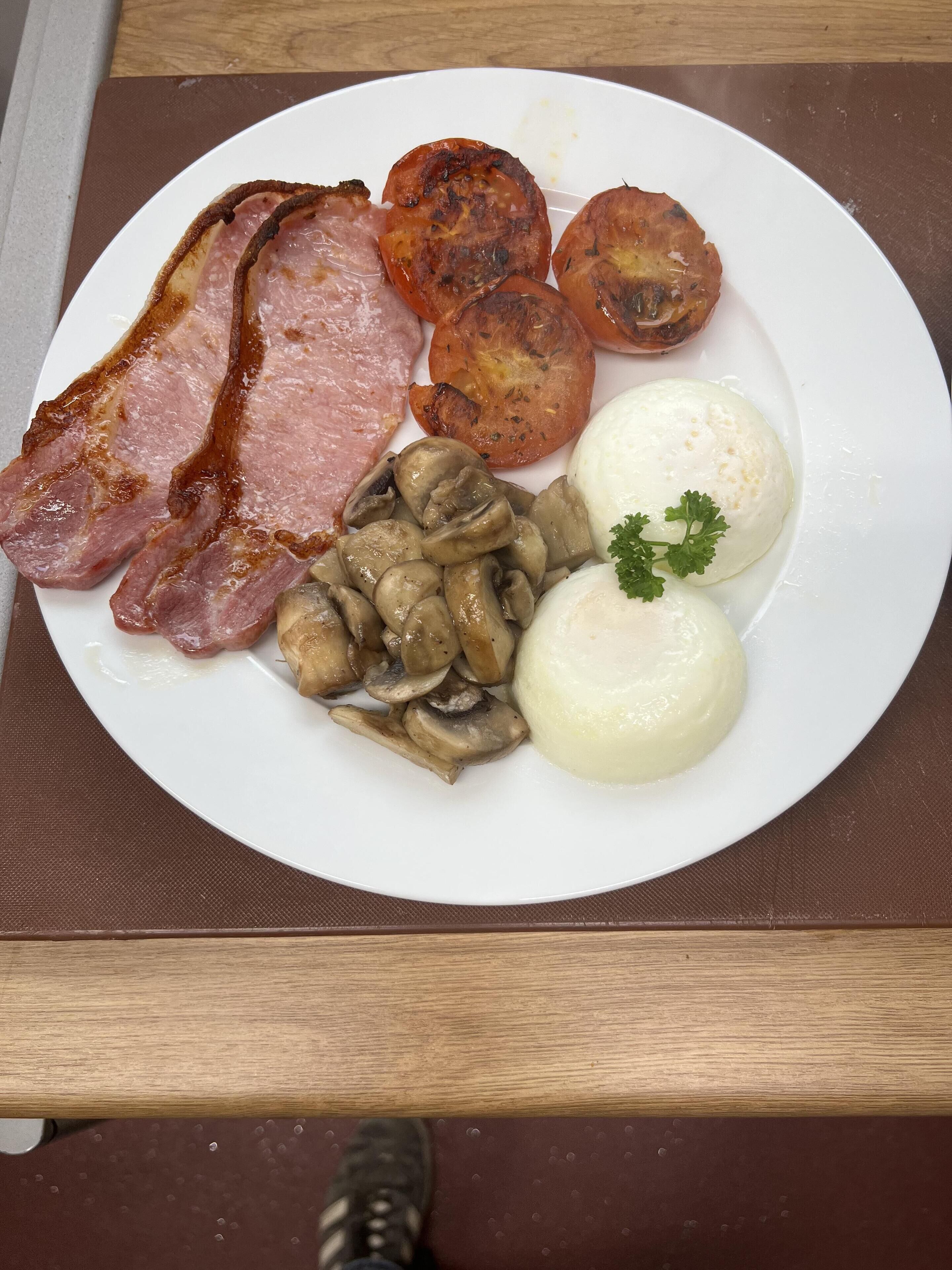Une assiette contenant des tranches de jambon, des tomates grillées, des champignons et des œufs durs, posée sur une table en bois.