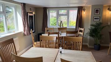 Un comedor con mesa y sillas de madera, ventanas grandes con cortinas, una planta en maceta y luces fijadas a la pared.