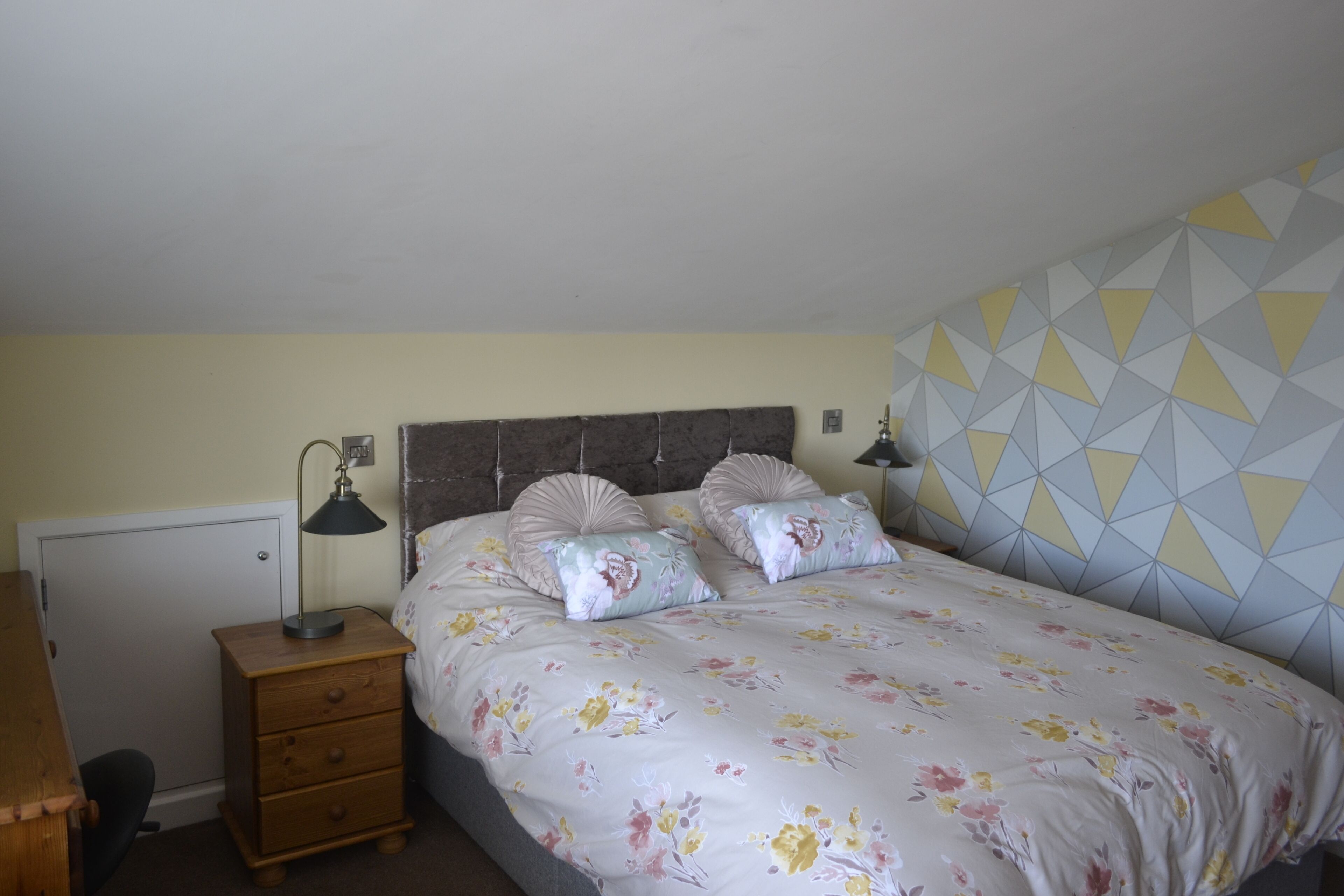 Une chambre à coucher comprenant un lit, deux lampes de chevet, une table de chevet en bois avec des tiroirs, un bureau et un mur décoré d’un motif.