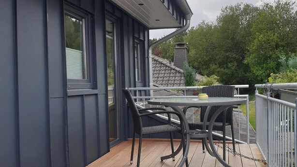 Una terraza de madera con una mesa y sillas, baranda metálica y vista a una casa con chimenea.