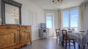 A dining area with a table set for two, a television on a stand, and a large window with curtains. - Cottage in Tharon: 2 bedrooms, terrace, parking, near the sea (Saint-Michel-Chef-Chef)