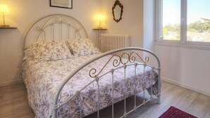 A bed with a floral bedspread, a radiator, a framed picture on the wall, and a window with a view of the outside. - Cottage in Tharon: 2 bedrooms, terrace, parking, near the sea (Saint-Michel-Chef-Chef)