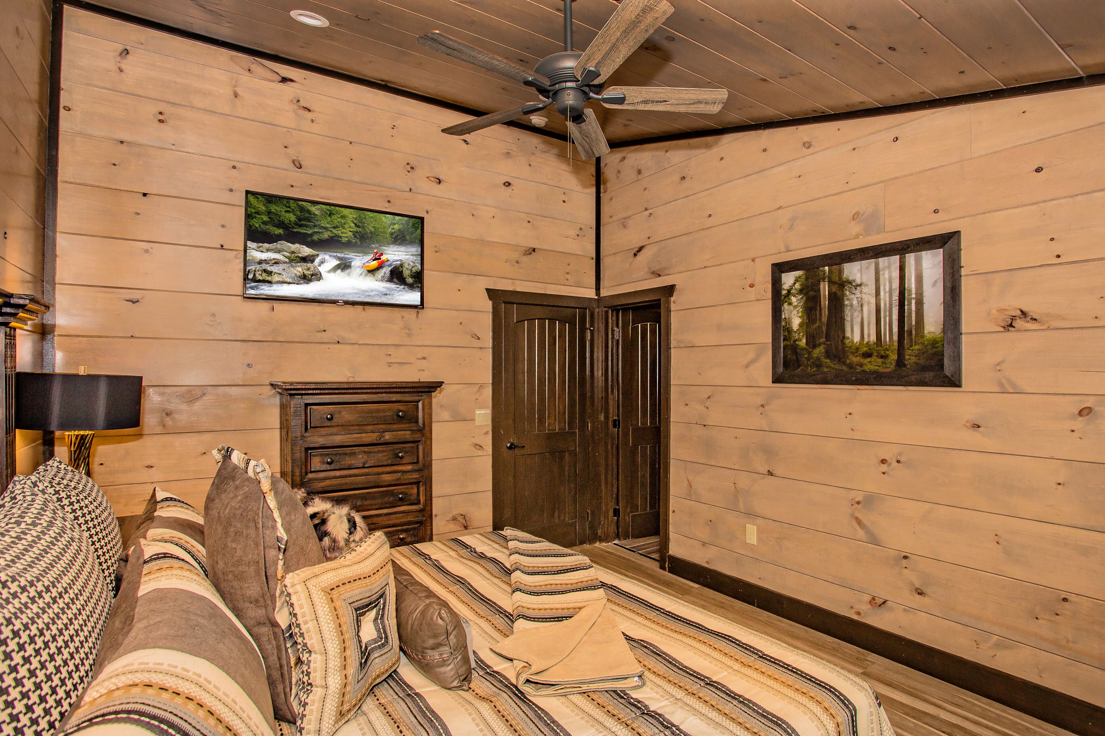 A wooden-paneled room with a bed, a ceiling fan, a television, and a door.