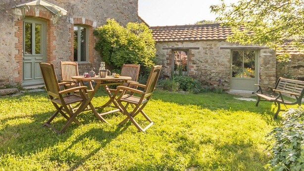 Un bâtiment en pierre avec un toit en tuiles, un petit jardin avec une table et des chaises, et un banc.