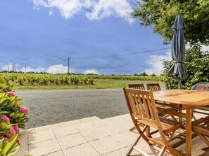 Eine Terrasse im Freien mit einem Holztisch und Stühlen, einem grauen Sonnenschirm und Blick auf einen Weinberg.