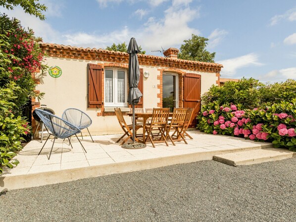 Ein kleines Haus mit Ziegeldach, einer Terrasse mit Tisch und Stühlen sowie ein Garten mit rosa Blumen.