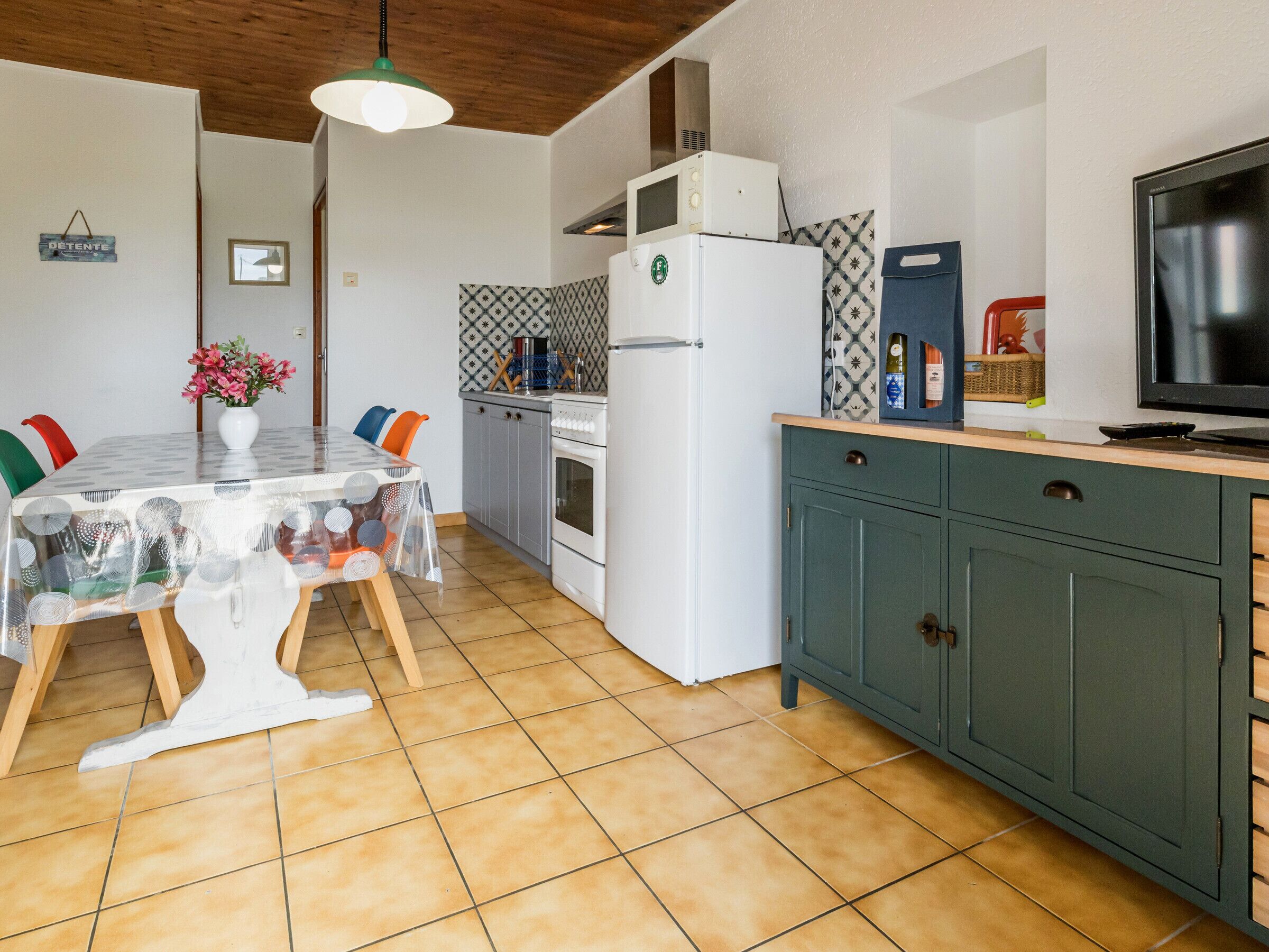 Eine Küche mit Fliesenboden, einem weißen Kühlschrank, einer Mikrowelle, einem Herd, einem Esstisch mit Stühlen und einem Fernseher auf einem Schrank.