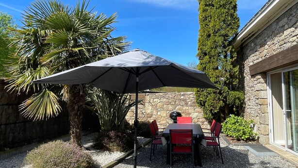 An outdoor patio with a table and chairs under a large umbrella, surrounded by plants and a stone wall. - Charming Cottage with Fireplace, Mezzanine, and Garden near Guérande (Saint-Molf)