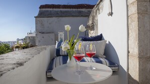 A rooftop terrace with a white table, two glasses of red wine, white flowers, and blue-striped cushions.