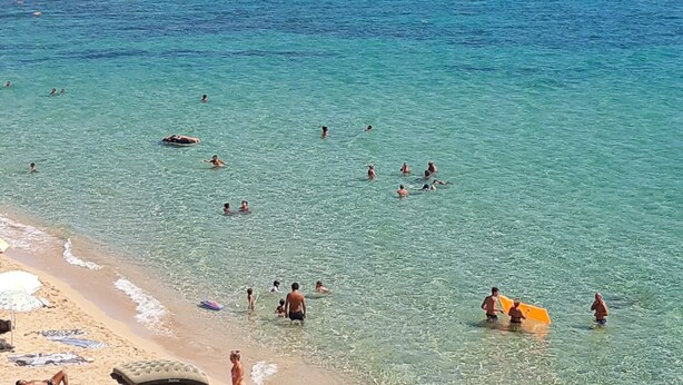 A beach with clear turquoise water, people swimming and sunbathing, and a sandy shore with umbrellas.