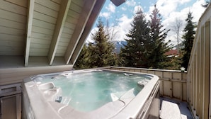 Ein Whirlpool auf einer Holzterrasse mit Blick auf hohe Nadelbäume und Berge im Hintergrund.