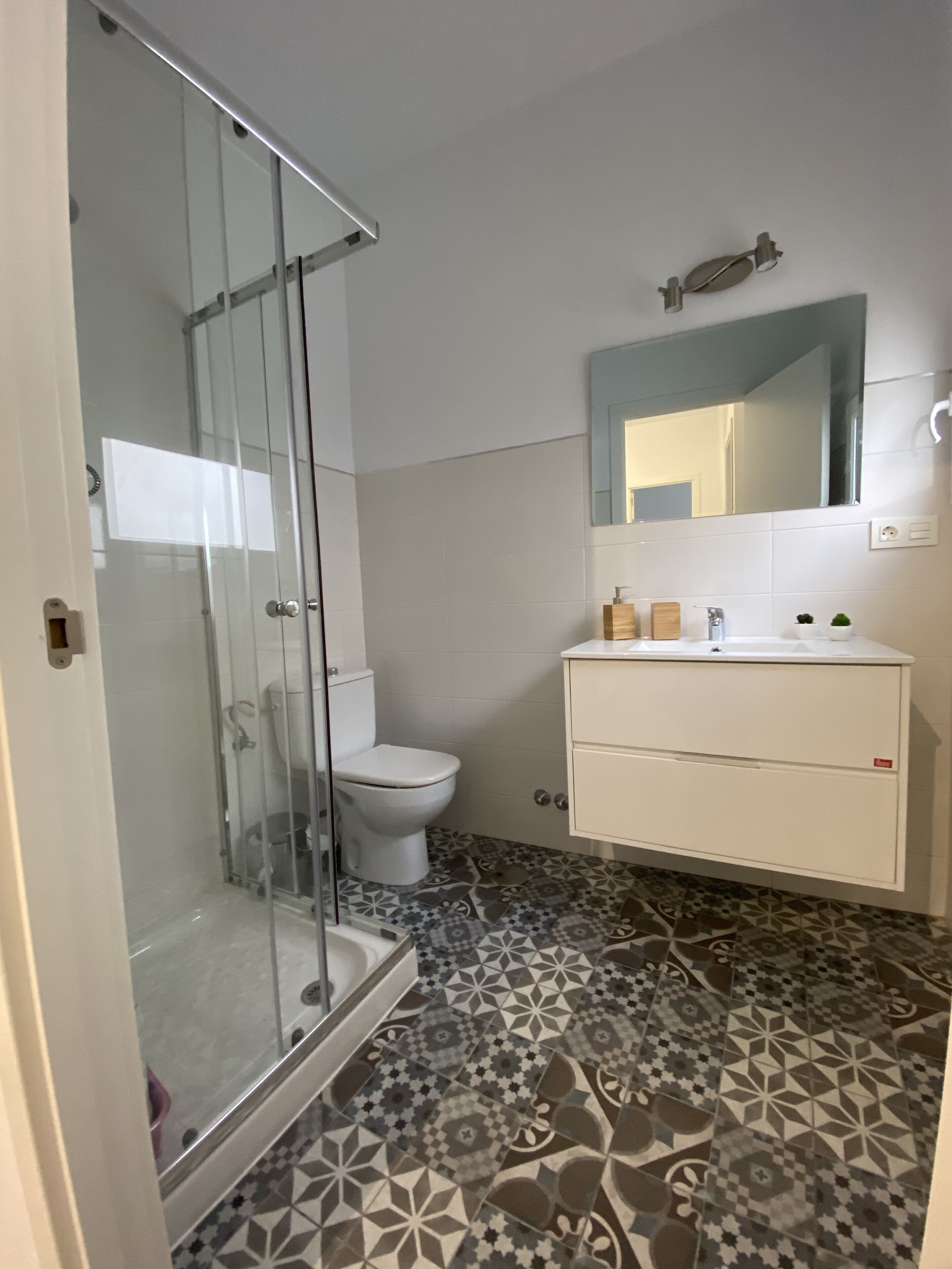 Un baño con cabina de ducha de vidrio, inodoro, lavamanos con espejo y baldosas de suelo estampadas.