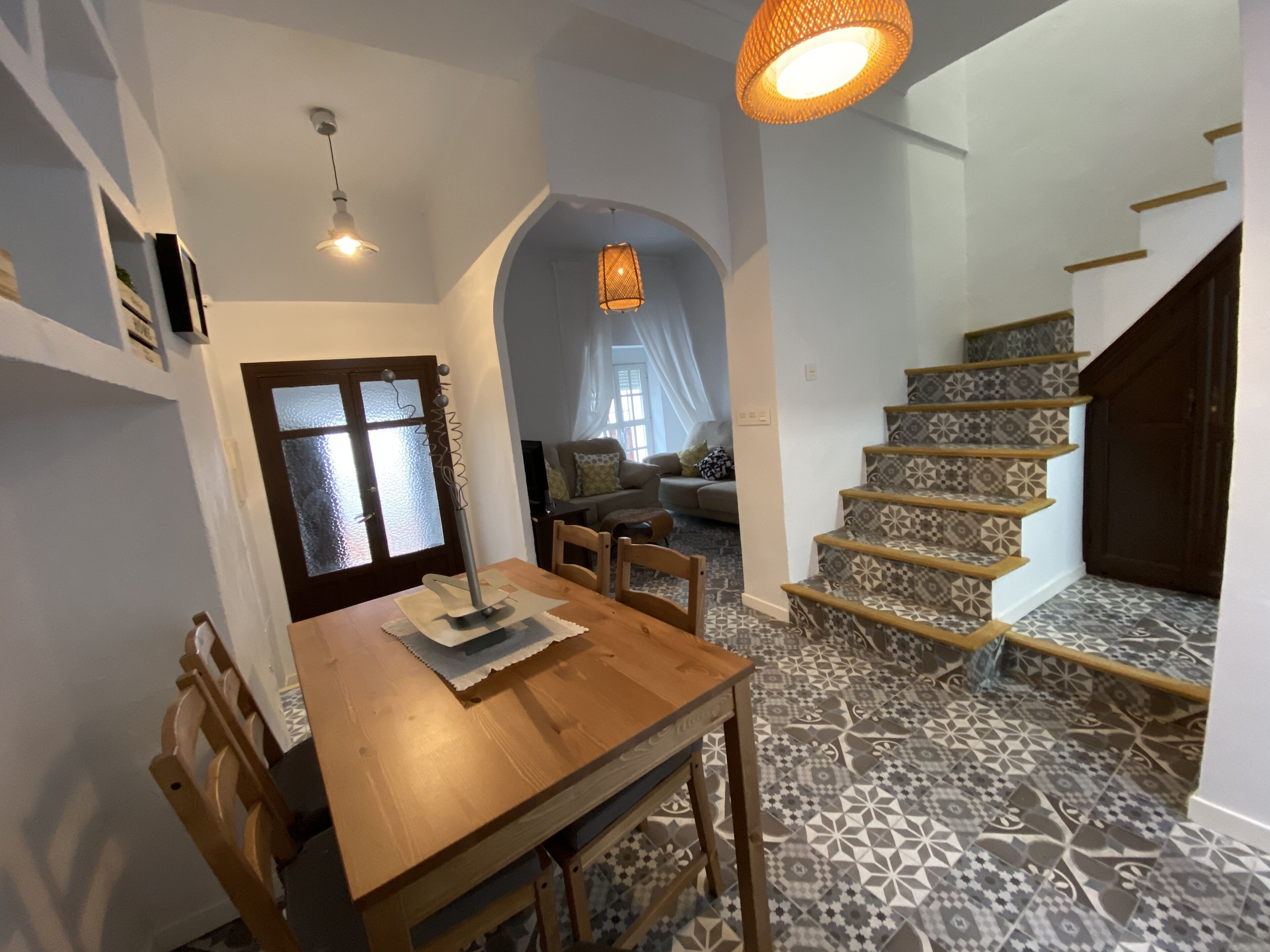 Un comedor con una mesa y sillas de madera, una escalera con baldosas estampadas y una lámpara colgante.