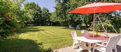A patio area with a table and chairs under a red umbrella, surrounded by a well-maintained lawn and trees.
