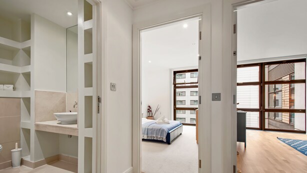 Une salle de bain moderne avec un lavabo blanc, des étagères et un miroir. Une chambre à coucher avec un lit et une vue sur un immeuble est visible à travers une porte ouverte.
