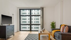 Un salon moderne doté d’un téléviseur à écran plat, d’une table basse en bois, d’un canapé gris et d’une grande fenêtre offrant une vue sur la ville.