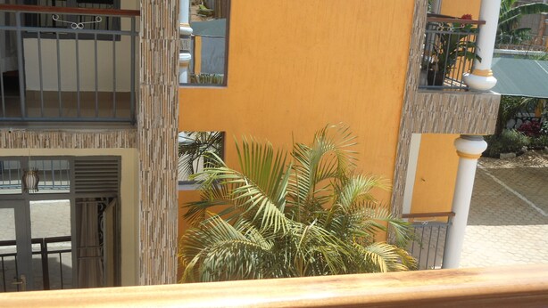 A building with a yellow facade, a balcony with a metal railing, and a potted plant in the foreground. - Glads Apartment (Kigali)